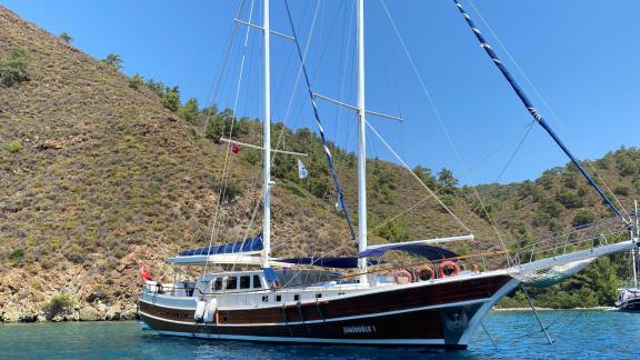 The Gulet Sahinoğlu, an impressive sailing ship with 7 cabins, lies quietly in a picturesque bay, surrounded by green hi